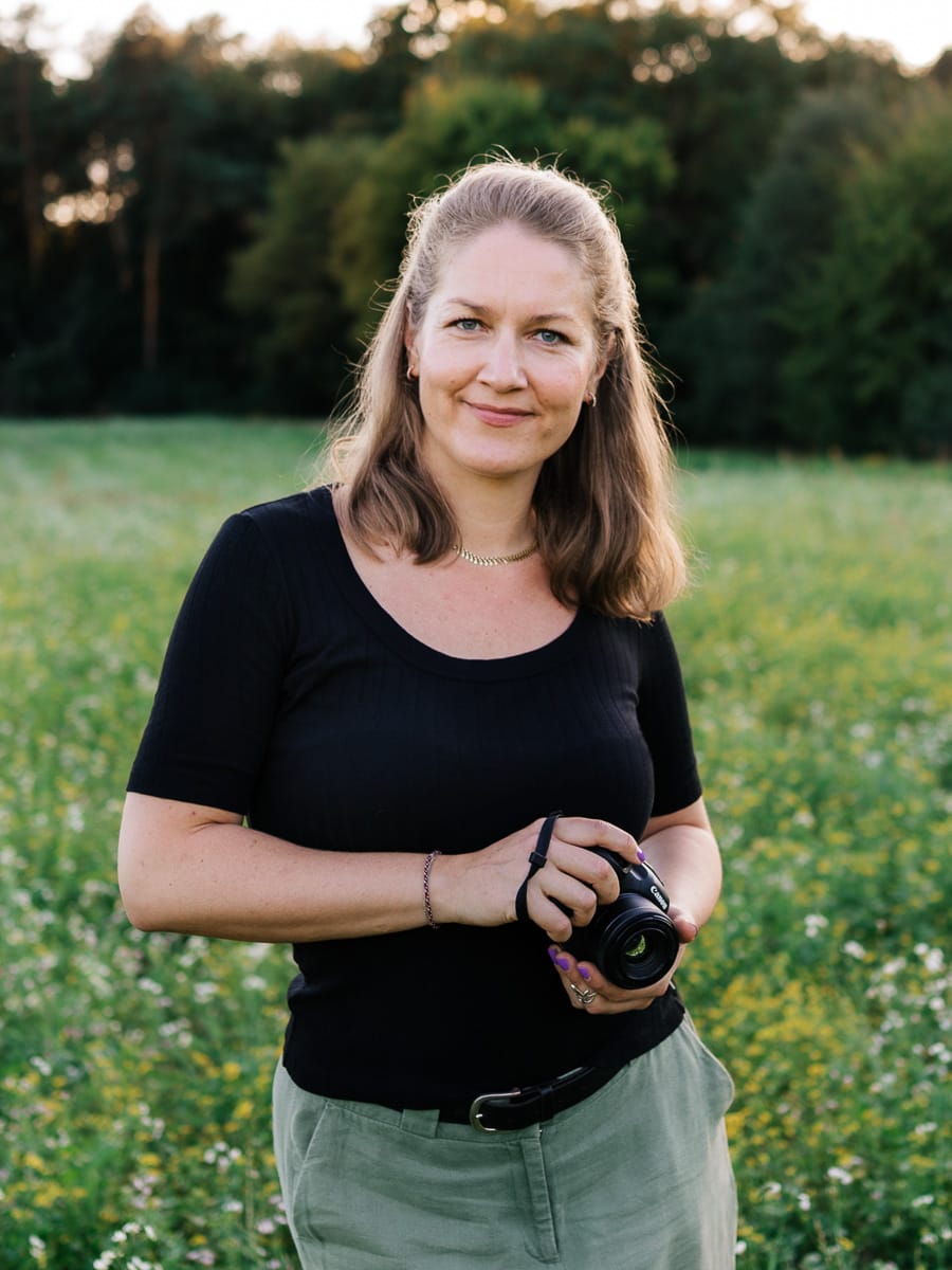 Hochzeitsfotograf Nora Peisger steht auf Wiese und hält Kamera in der Hand und lächelt